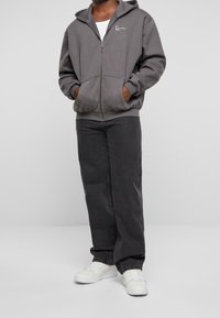 Sudadera gris con cremallera y bolsillo tipo canguro, combinada con jeans sueltos de color gris oscuro y zapatillas blancas. La sudadera tiene un logo en el pecho.