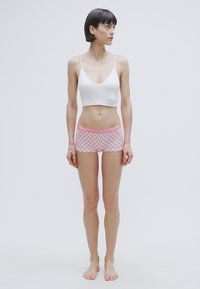 Λευκό μπουστάκι με λεπτές τιράντες συνδυασμένο με ροζ κοντό παντελόνι με σχέδια και διακοσμητική ζώνη στη μέση. Το μοντέλο στέκεται ξυπόλυτο σε ουδέτερο φόντο.