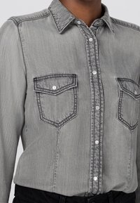 Gray denim shirt with a pointed collar, two chest pockets, and button-front closure. Features contrast stitching and a soft texture.