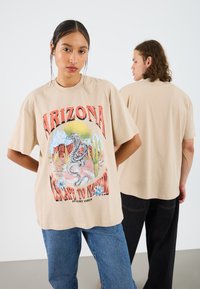 Beige oversized t-shirt featuring a graphic print of a snake, desert landscape, and text. Fabric is soft cotton with a relaxed fit.