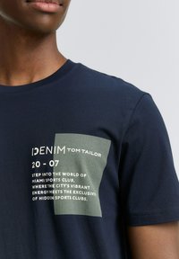 Navy modra bombažna majica z teksturiranim zelenim grafičnim tiskom in belim besedilom na zgornji levi strani. Klasičen kroj s krožnim ovratnikom.
