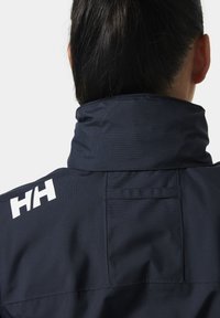 Marineblauw jack met een hoge kraag, gestructureerd materiaal en een wit logo. Voorzien van een achterzak en gestikte details.