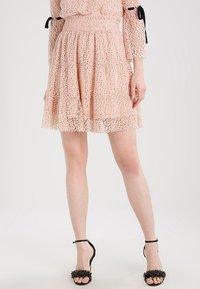 Spetskjol i mjuk rosa med ett lagerdessin. Har ett smockat midjeband och en intrikat spetsmönster. Bärs med svarta klacksandaler.