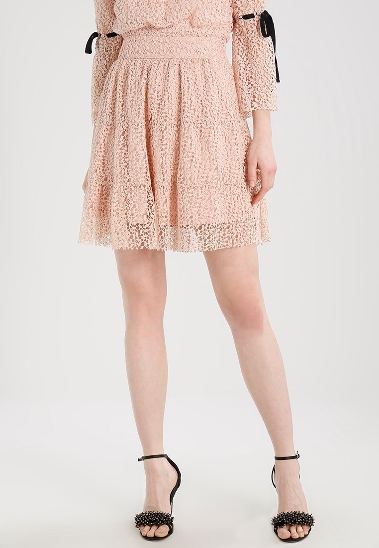 Spetskjol i mjuk rosa med ett lagerdessin. Har ett smockat midjeband och en intrikat spetsmönster. Bärs med svarta klacksandaler.