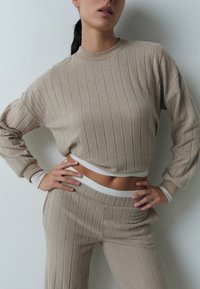 Frau trägt beigen gerippten Strickpullover und passende Hose, Hände in die Hüften gestützt, auf Bauchnabelhöhe beschnitten, steht vor einer weißen Wand.