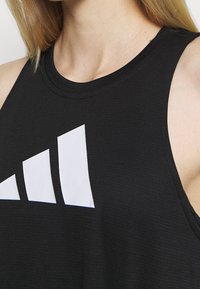 Gros plan d'une personne portant un débardeur noir avec un grand logo Adidas à trois bandes blanches sur la poitrine.