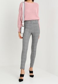 Roze gebreide trui, grijze gebloemde hoge taille broek met riemdetail, strakke pasvorm, en zwarte hakken met bandjes. Witte schoudertas.