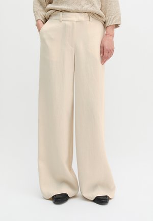 Beżowe spodnie o szerokich nogawkach noszone z rękami w kieszeniach, zestawione z beżowym swetrem dzianinowym i czarnymi butami o kwadratowym nosku.