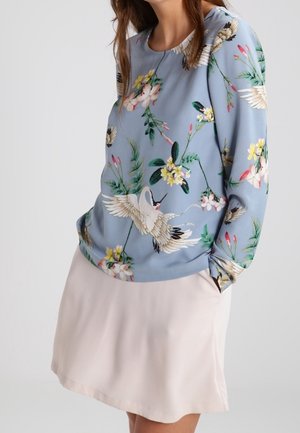 Niebieska bluzka z długim rękawem z białym nadrukiem żurawi i kwiatów, zestawiona z jasnobeżową spódnicą, ręce w kieszeniach, na gładkim tle.