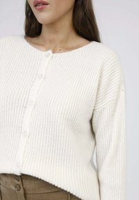 Cardigan en tricot de couleur crème avec un col rond, doté d'une fermeture à boutons et d'une texture côtelée, assorti d'une ceinture en daim marron.