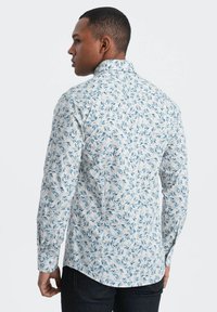 Lange mouwen shirt van lichte stof met een blauw bladenpatroon op een witte achtergrond. Heeft een puntige kraag en knoopmanchetten.