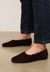 Ballerines en daim marron avec un embout rond et un bord volé. Les semelles sont plates et la texture semble douce et lisse.