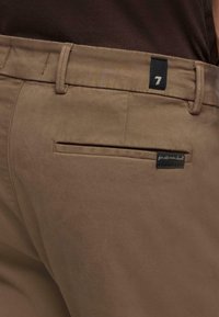 Bruine broek gemaakt van gladde stof. Heeft een achterzak met een zwart label met het cijfer "7" en een ander label met de tekst "for all mankind."