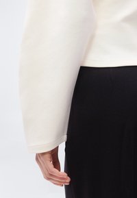 Ελαφρύ κρεμ φούτερ με φαρδύ μανίκι και κυρτή ραφή, σε συνδυασμό με μαύρο κάτω μέρος. Μαλακή υφή υφάσματος, με ελάχιστη ορατή ραφή.