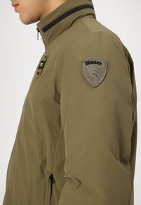 Veste vert olive avec un col montant, fermeture éclair, et un patch Blauer sur l'épaule gauche. Tissu lisse avec un fini mat.