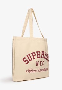 Sac fourre-tout en toile beige avec deux poignées, portant un texte bordeaux qui indique "SUPERDRY N.Y.C. Essentiels Athlétiques." Design simple, matériau robuste.