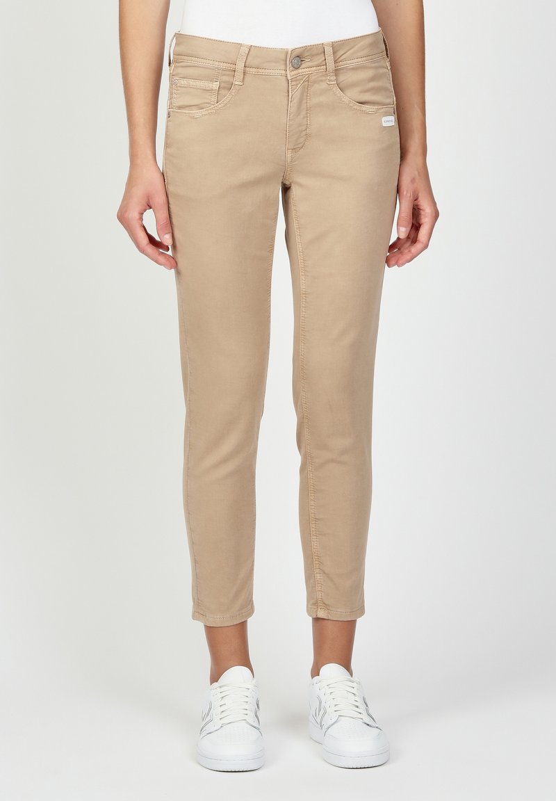 Personne portant un pantalon beige ajusté et des baskets blanches, debout devant un fond uni clair, les mains détendues le long du corps.
