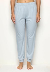 Mujer con pantalones jogger de cintura elástica azul claro y tobillos ajustados, de pie descalza frente a un fondo blanco liso.