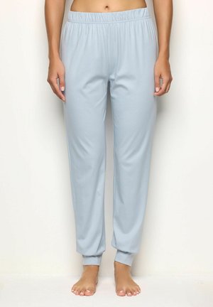 Mujer con pantalones jogger de cintura elástica azul claro y tobillos ajustados, de pie descalza frente a un fondo blanco liso.