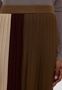 Jupe en maille plissée avec des blocs de couleur verticaux beige, bordeaux et marron, portée avec un haut à manches longues taupe.