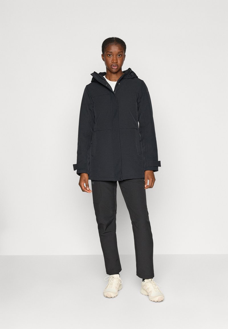 Veste imperméable noire avec capuche, comportant deux poches avant et une fermeture éclair. Portée avec un pantalon noir et des baskets de couleur claire.