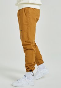 Cargo-byxor i senapsgul tyg, med elastiska fållar, sidofickor och en strukturerad yta. Matchas med vita Nike-sneakers.