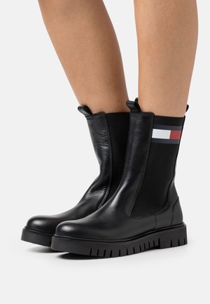 Czarne skórzane botki ze sprężystymi panelami po bokach, ozdobione paskowanym akcentem u góry i solidną gumową podeszwą. Gładka faktura, nowoczesny design.