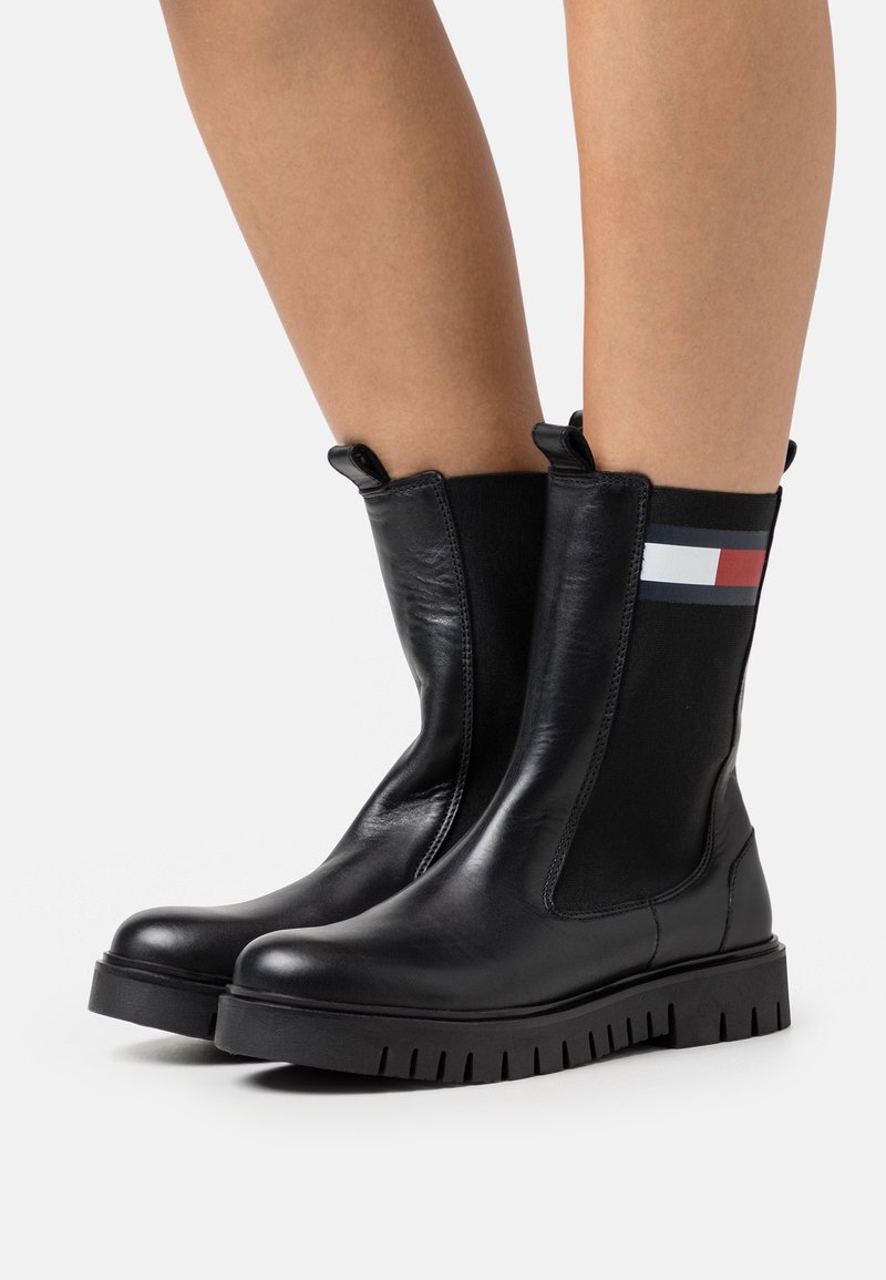 Bottes chelsea en cuir noir avec panneaux latéraux élastiques, ornées d'un accent rayé en haut et d'une semelle en caoutchouc robuste. Texture lisse, design moderne.