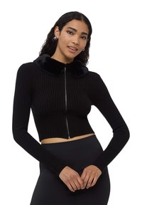 Czarny prążkowany krótki kardigan z długimi rękawami, z zamkiem błyskawicznym i sztucznym futerkiem przy kołnierzu. Gładka textura, minimalistyczny design.