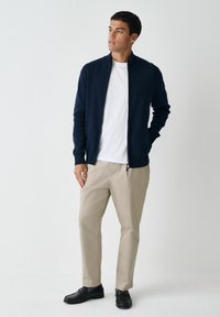 Navy zip-up jacket made of knit material, paired with a white t-shirt, beige trousers, and black loafers, standing against a plain white background.