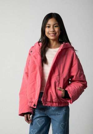 Uśmiechnięta dziewczyna z długimi ciemnymi włosami, ubrana w jasnoróżową pikowaną kurtkę, jasną koszulę w paski oraz niebieskie dżinsy z wzorem serc, stojąca na szarym tle.