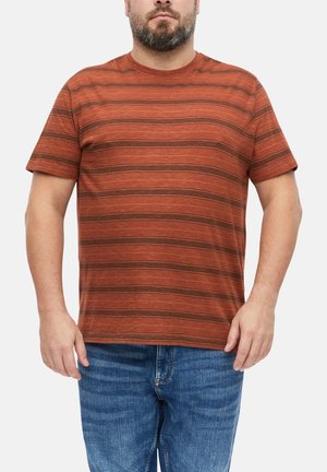 Mężczyzna w rdzawej, krótkiej, pasiastym T-shircie i niebieskich dżinsach, stojący na jednolitym jasnym tle.