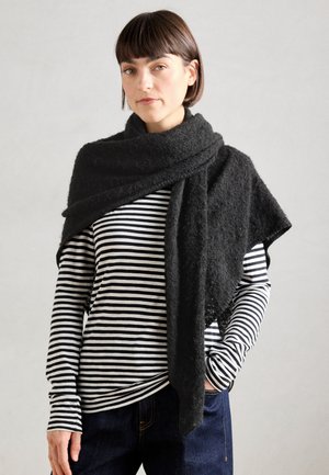 Femme aux cheveux bruns courts portant un châle texturé noir sur un chemisier à manches longues à rayures noires et blanches et un pantalon foncé, debout devant un fond gris.
