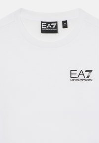 T-shirt bianco in cotone con collo rotondo. Presenta un logo nero EA7 Emporio Armani nella zona sinistra del petto. Texture morbida e liscia.