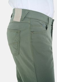 Olive green pants made of cotton, featuring a smooth texture, standard pockets, and a simple design with a leather patch on the waistband.