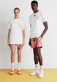 Camisetas brancas com um pequeno design de sol, combinadas com calções curtos coloridos e estampados, apresentando elementos em rosa, azul e florais. Calçado laranja brilhante e branco.