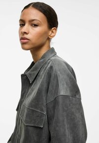 Szara zamszowa kurtka z kołnierzem, dwiema przednimi kieszeniami i luźnym krojem. Miękka tekstura i matowe wykończenie, podkreślające szczegółowe przeszycia.