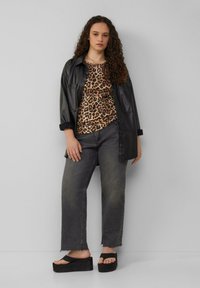 Schwarze Lederübershirt, das über einem Leopardenmuster-Oberteil getragen wird, kombiniert mit grauen, weit geschnittenen Jeans und schwarzen Plateausandalen mit strukturierter Oberfläche.