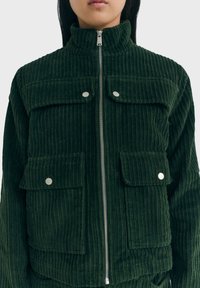 Groen ribfluwelen jack met een hoge kraag, zilveren rits en twee opgestikte zakken aan de voorkant. Heeft een geribbelde textuur en knoopdetails.