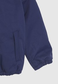Námořnická modrá bunda s elastickými manžetami, postranní kapsou a hladkou texturou látky. Jednoduchý design bez dalších vzorů nebo akcentů.