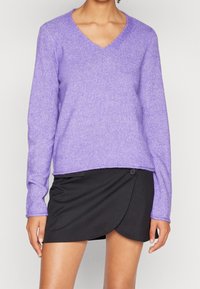 Pull en V violet en matière douce, avec des manches longues et une coupe décontractée, associé à une jupe portefeuille noire avec un détail de bouton.