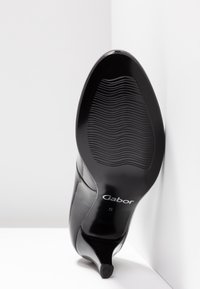 Chaussure à talons hauts noirs avec une finition brillante, dotée d'une semelle texturée ondulée et d'un bout pointu, étiquetée « Gabor » sur le côté.