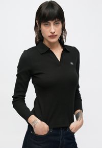 Polo a maniche lunghe nera con una texture a coste, caratterizzata da un colletto e un piccolo logo marchiato sul lato sinistro.