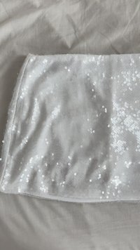 Tessuto bianco coperto di piccole paillettes lucide disteso su una superficie beige testurizzata, con leggere pieghe e grinze.