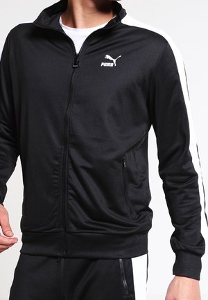 Homme portant une veste de survêtement noire Puma avec des bandes blanches sur les manches et des poches zippées, associée à un pantalon assorti.