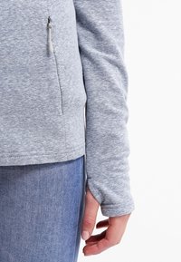 Felpa grigia chiara a maniche lunghe con una texture liscia, dotata di una tasca con zip sul lato e un orlo curvo. Indossata con jeans blu.