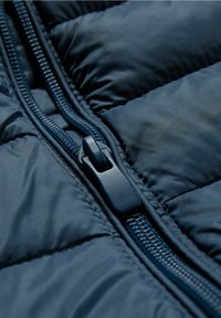 Section de doudoune bleu marine foncé avec des panneaux matelassés et une fermeture éclair proéminente. La fermeture éclair comporte une tirette texturée et un rail lisse.