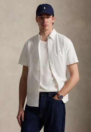 Jeune homme portant une casquette bleu marine, une chemise blanche à manches courtes boutonnée sur un tee-shirt blanc, un jean foncé et une montre-bracelet avec un bracelet en cuir.
