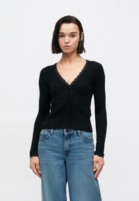 Μαύρο ριμπ πλεκτό ζακέτα με δαντέλα στο τελείωμα, με V-λαιμόκοψη και μακριά μανίκια, συνδυασμένο με ψηλόμεσα μπ λε τζιν. Διακριτικό κούμπωμα με κουμπιά.