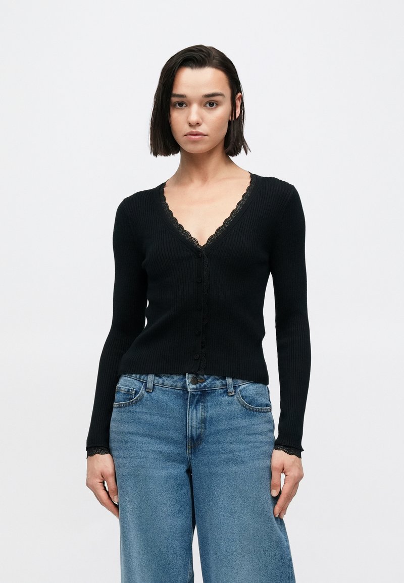 Μαύρο ριμπ πλεκτό ζακέτα με δαντέλα στο τελείωμα, με V-λαιμόκοψη και μακριά μανίκια, συνδυασμένο με ψηλόμεσα μπλε τζιν. Διακριτικό κούμπωμα με κουμπιά.